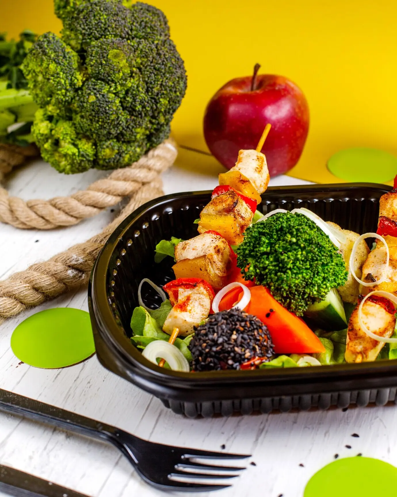 Vista lateral de brochetas de pollo frito con verduras frescas (brócoli y cebollas) en una caja de entrega.